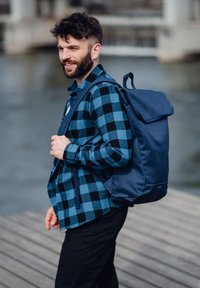 Dunkelblauer Rucksack mit Klappenverschluss und gepolsterten Trägern, getragen über einem schwarz-blau karrierten Hemd, vor einem Wasserhintergrund.