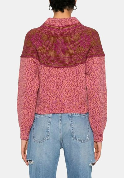 Donna che indossa un maglione lavorato a maglia con motivo rosa e marrone e jeans azzurri strappati, vista da dietro su uno sfondo semplice.