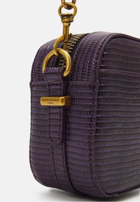 Lila texturerad läderväska med dragkedjestängning, guldiga detaljer och en märkestagg. Har en rundad form och synliga stygndekorationer.