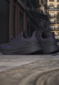 Par mørkelilla og sorte Adidas Ultraboost løbesko placeret på en bygade med bygninger i baggrunden.