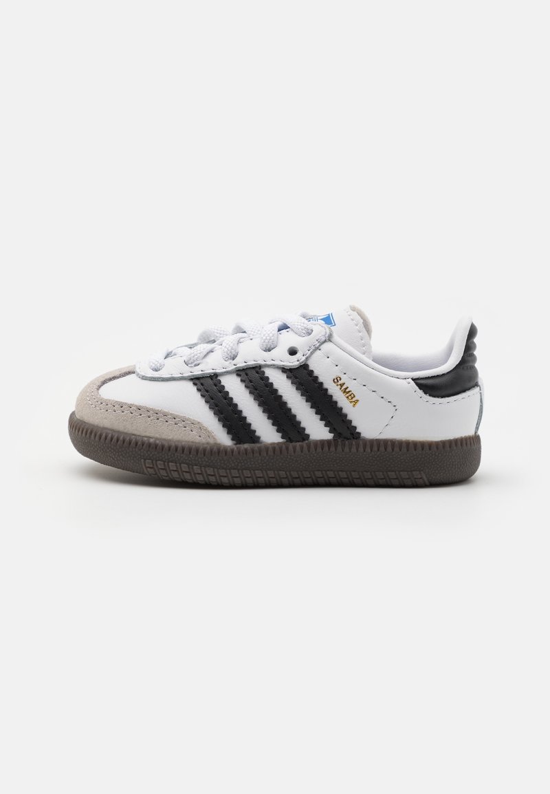 White toddler sneakers with black stripes, suede toe cap, textured rubber sole, and blue tongue label. Lightweight, sporty design.