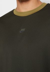 Camiseta atlética de color verde oscuro con un cuello acanalado de color verde claro, que presenta un sutil logo de Nike y una tela texturizada con pequeños agujeros de ventilación.