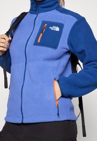 Polarowa kurtka w jasnoniebieskim i ciemnoniebieskim kolorze, z zamykaną na zamek kieszenią na piersi z pomarańczowymi akcentami i czarnym zamkiem oraz wysokim kołnierzem.