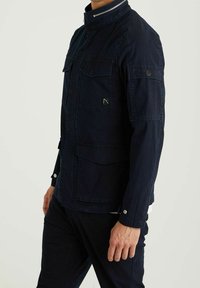 Navy blue jacket with a high collar, multiple front pockets, contrasting stitching, silver hardware accents, and a smooth, textured finish.