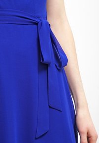 Kungsblå klänning med en mjuk textur, prydd med ett knytband av satin i midjan som skapar en flytande silhuett.