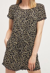 Czarny kombinezon w kwiaty z małymi żółtymi i białymi wzorami, krótkimi rękawami, elastycznym pasem i koronkowym wykończeniem na rękawach oraz u dołu.