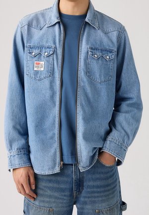 Mand iført en lyseblå denim lynlåstrøje over en blå t-shirt og blå denim bukser med lommer, hånd i lommen.