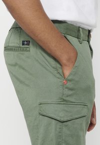 Grüne Baumwoll-Cargo-Hosen mit seitlichen Taschen, einer Gesäßtasche und einem kleinen Logo-Tag mit roten Akzenten und Knopfdetails.