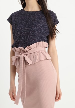 Vrouw draagt een marineblauw gespetterd kort mouwtopje, ingestopt in een hoog getailleerde, roze geribbelde rok met een gerimpelde riem die in een strik is gebonden.