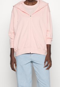 Osoba nosící světle růžovou mikinu na zip přes světle růžové tričko Nike a světle modré džínové kalhoty, stojící s uvolněnými pažemi.