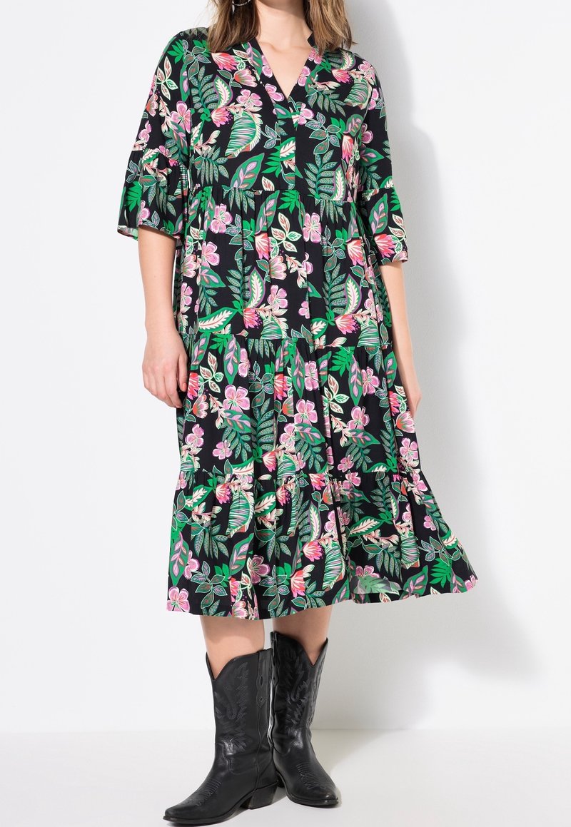 Robe noire avec des motifs floraux et de feuilles en rose et vert. Dotée d'un col en V, de manches trois-quarts et d'un ourlet en couches. Portée avec des bottes de cow-boy noires.