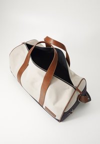Sac de voyage en toile avec des accents en cuir marron, fermeture zippée sur le haut, corps beige et base bleu foncé, doté de poignées doubles pour le transport.