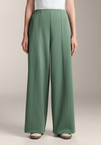Široke hlače iz mehkega zelene tkanine, ki se ponašajo z navpičnimi šivi in gladko teksturo, dopolnjene z visokim pasom.