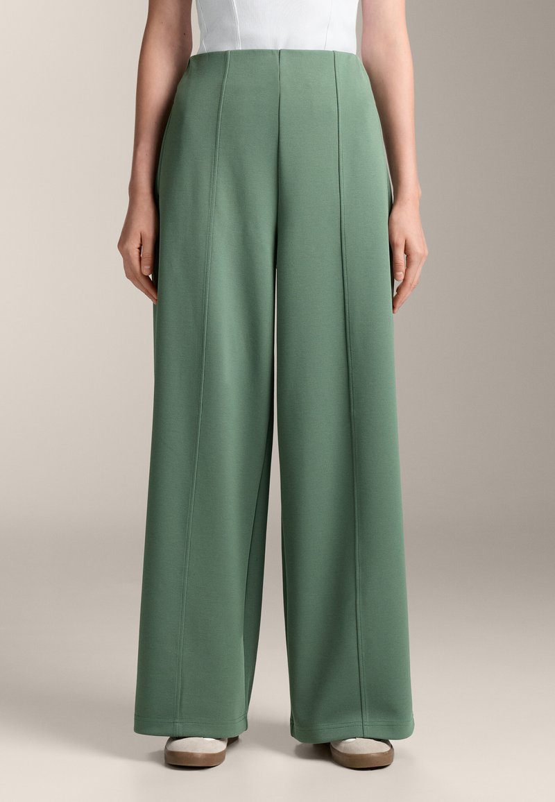 Široke hlače iz mehkega zelene tkanine, ki se ponašajo z navpičnimi šivi in gladko teksturo, dopolnjene z visokim pasom.