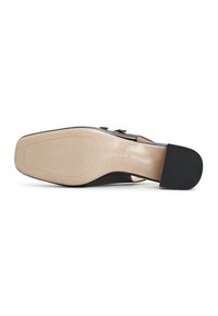 Zapato de tacón alto negro con punta afilada, que presenta una suela beige, parte superior negra texturizada y detalles decorativos en las correas.