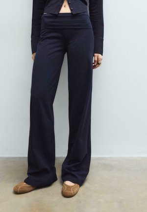 Personne portant un pantalon large noir taille haute et des chaussures marron à enfiler, debout sur un sol en béton lisse devant un mur blanc.