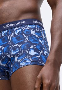 Közeli kép egy személyről, aki kék mintás Björn Borg boxeralsót visel, a törzs, derék és a comb felső része látható egy sima háttér előtt.