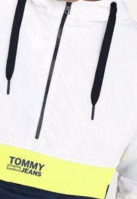 Vit huvtröja med dragkedja, med svarta dragsnören och ett gult rand med "TOMMY JEANS"-logotyp. Slät textur, sportig design.