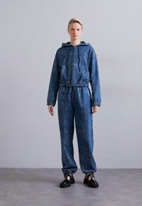 Îmbrăcăminte din denim formată dintr-o jachetă cu fermoar și glugă, dotată cu buzunare și talie elastică, asociată cu pantaloni cu manșete elastice asortate.