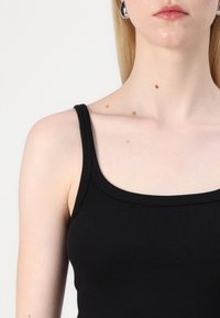 Svart ribbet tanktopp med en dyp utringning og brede stropper, som viser en tettsittende design og teksturert stoff. Justerbar passform for komfort.