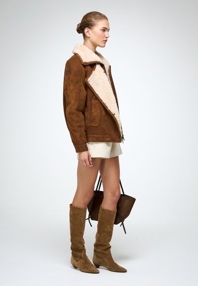 Jachetă din suede maro cu guler de blană crem, două buzunare frontale, asortată cu pantaloni scurți bej și cizme din suede maro până la genunchi. Geantă maro inclusă.