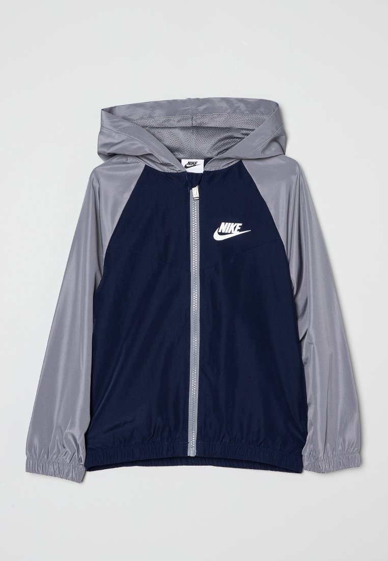 Veste coupe-vent marine et grise avec une capuche, fermeture éclair à l'avant, poignets élastiques et un logo Nike blanc sur la poitrine. Texture du tissu lisse.