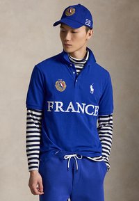 Joven con gorra azul y polo con la palabra "FRANCE" y emblema, sobre una camiseta de manga larga de rayas en blanco y negro.