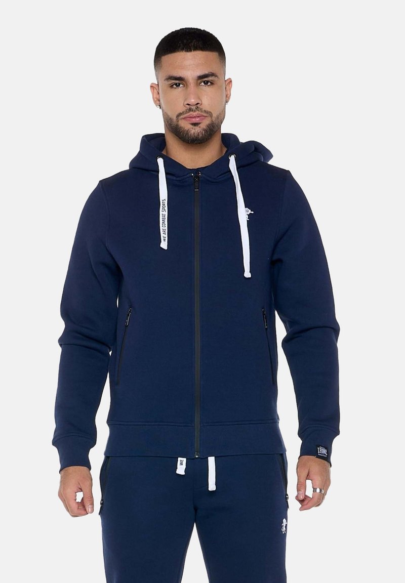 Homme portant un sweat à capuche bleu marine zippé avec des cordons blancs et un pantalon de survêtement bleu marine assorti sur un fond uni.
