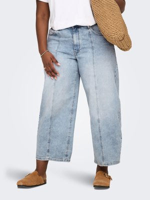 Osoba v svetlomodrých širokých džínsových nohaviciach skrátenej dĺžky, s opálenými slip-on topánkami, drží veľkú pletenú tašku hnedej farby, doplnená prsteňmi a náramkom.