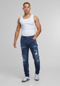 Jean en denim bleu ajusté avec des patchs déchirés, un léger dégradé et un accent de logo rouge. Assorti à un débardeur blanc et des baskets.
