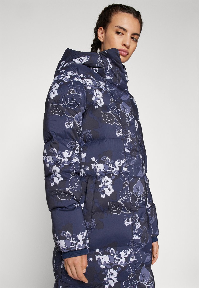 Doudoune bleu foncé avec motif floral et de feuilles blanches. Elle dispose d'un col haut, de poches latérales et d'une texture matelassée.