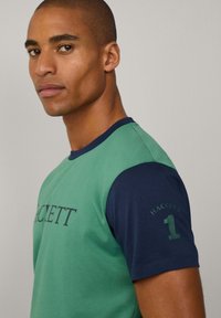 Camiseta de manga corta verde y azul marino con el logo "HACKETT" en el pecho y el número "1" en la manga. Fabricada en una suave mezcla de algodón, con un diseño deportivo.