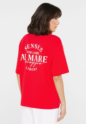 Woman with shoulder-length dark hair wearing a loose red T-shirt with white text on the back and white pants, facing sideways.