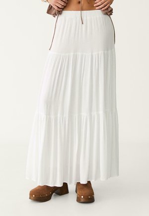 Femme portant une longue jupe blanche à volants avec des sabots marron compensés sur un fond uni.