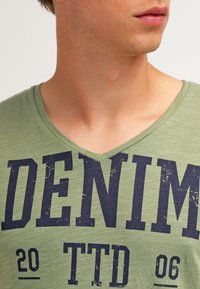 Olivově zelené tričko s výstřihem do V z texturované látky s velkým námořnickým modrým potiskem "DENIM 20 TTD 06". Casual design, krátké rukávy.