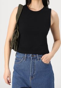 Black sleeveless top with a rounded neckline, paired with high-waisted blue denim jeans. Model carries an olive green bag with zippers.