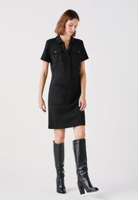 Schwarzes Hemdkleid mit kurzen Ärmeln, Knopfleiste und zwei aufgesetzten Taschen; kombiniert mit knielangen schwarzen Lederstiefeln mit blockigem Absatz.