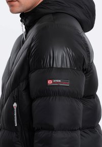 Schwarze Puffjacke mit glänzender Oberfläche, ausgestattet mit Kapuze, gestepptem Design und einem Logopatch am linken Ärmel. Seitlicher Reißverschlussbereich sichtbar.