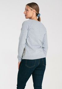 Jasnoszary sweatshirt z teksturowanym wzorem po bokach i długimi rękawami, zestawiony z ciemnoniebieskimi dżinsami. Widok z tyłu ukazuje luźny krój.