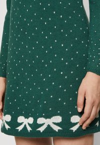 Grön stickad klänning med en texturerad mönster av vita v-formade motiv och vita rosetter i fållen, med långa ärmar och figursydd design.