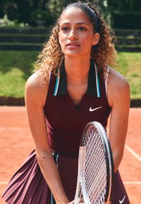 Robe de tennis bordeaux avec jupe plissée et col noir, dotée d'accents turquoise et d'un logo Nike blanc, tenue avec une raquette de tennis.