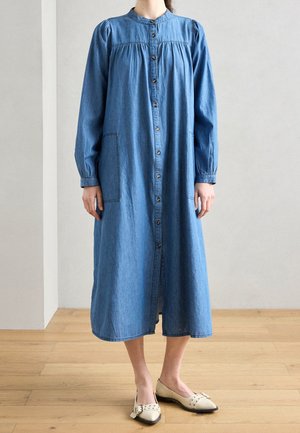 Woman wearing a long-sleeve, button-down blue denim dress with patch pockets and beige pointed-toe flats standing on a wooden floor.