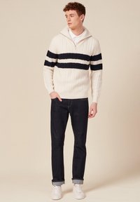 Weißer Kabelstrickpullover mit marineblauen Streifen, Reißverschluss-Kragen, dunkler Jeans und weißen Sneakern. Das Design hebt die Textur und kontrastierende Farben hervor.