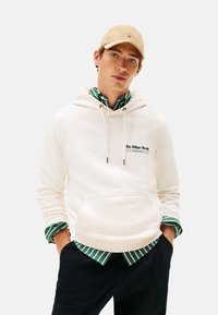 Joven con gorra beige, sudadera blanca con texto en el pecho, camisa a rayas verde debajo y pantalones oscuros, de pie con las manos en los bolsillos.