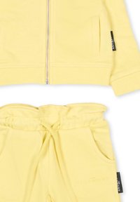 Żółta bluza z kapturem z przednim zamkiem i ściągaczami na rękawach, w zestawie z dopasowanymi żółtymi dresami z elastyczną talią i bocznymi kieszeniami.