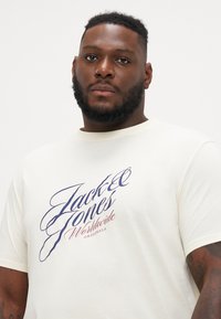 Krema pamučna majica s velikim plavim i crvenim stiliziranim natpisom "Jack & Jones Worldwide Originals", kratkim rukavima i okruglim izrezom.