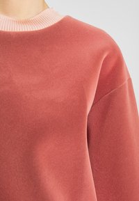 Sudadera coral suave con un cuello de canalé rosa; presenta una textura lisa y un ajuste relajado. Diseño sin costuras en los hombros para mayor comodidad.