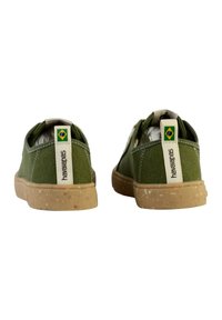 Des chaussures en toile vertes avec une semelle en caoutchouc. Elles présentent une languette à l'arrière et une étiquette de marque avec le drapeau brésilien. Surface texturée et forme arrondie.