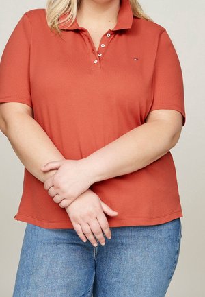 Osoba ubrana w polo koloru rdzy z trzema rozpiętymi guzikami, skrzyżowanymi rękami i niebieskimi dżinsami na jednolitym tle.