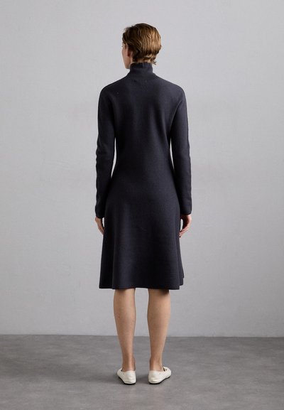 Dunkelgraues, langärmliges Kleid mit hohem Kragen, aus weichem Stoff, tailliert und bis zur Mitte der Wade schwingend. Getragen mit flachen weißen Schuhen.